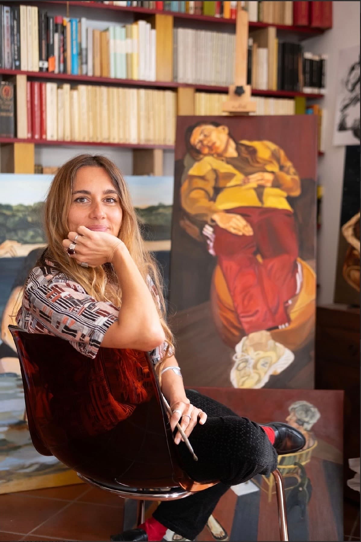 Daniela sits beside a figurative painting in her studio, with books on shelves behind her.