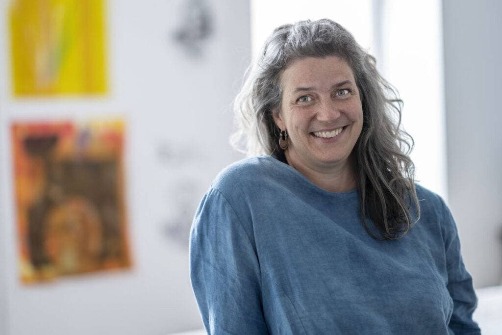 Leslie Menagh smiles in a blue linen top, with long grey hair, in a bright room with colourful prints on the wall behind her.