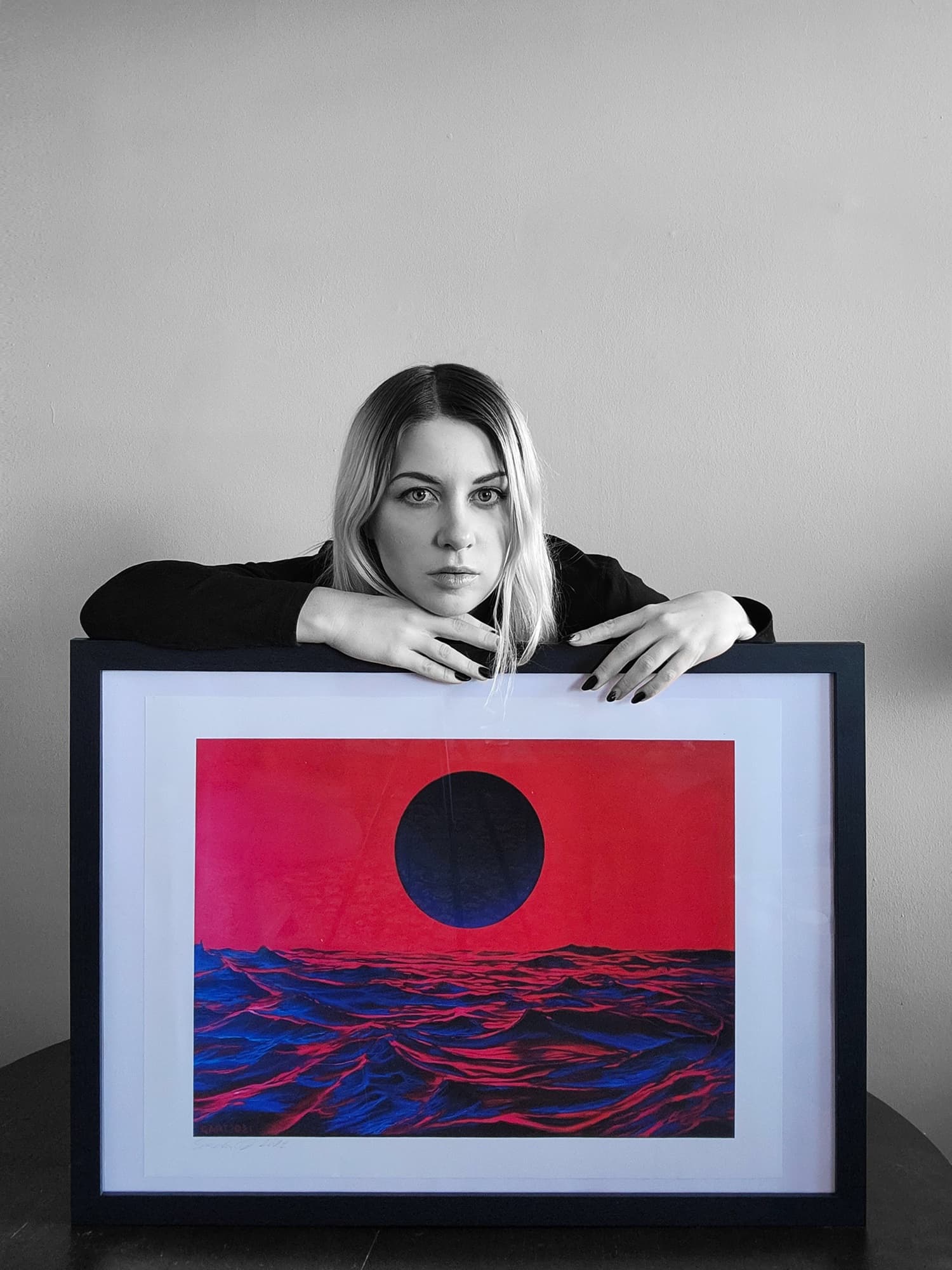 Sasha rests her arms on a framed print of a dark sun over a red and blue sea.